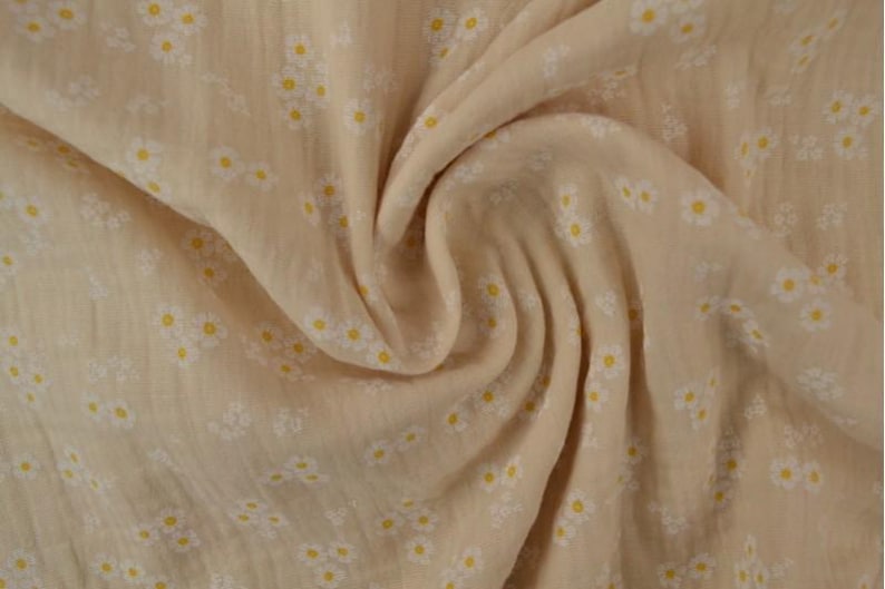 May include: Close-up of a beige fabric with a daisy pattern. The fabric is soft and flowing, with small white daisies and yellow centers scattered across the surface. The fabric is gathered in soft folds.