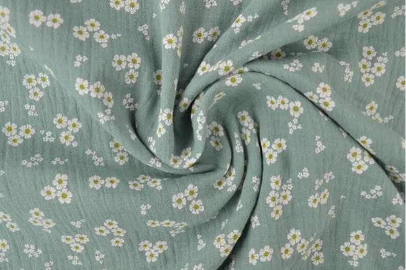 May include: A close-up of a soft, sage green fabric with a delicate floral pattern. The fabric is covered in small white flowers with yellow centers, creating a charming and airy design. The fabric is draped, showing its texture.