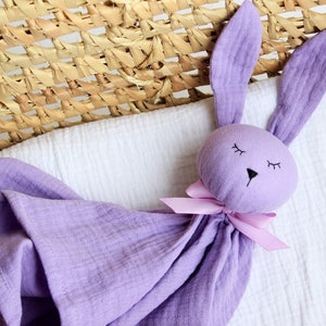 K&ouml;nnte beinhalten: Eine lavendelfarbene, bunnyf&ouml;rmige Sicherheitsdecke mit einer rosa Schleife. Die Decke ist aus weichem, strukturiertem Stoff und hat ein gesticktes Gesicht mit geschlossenen Augen.
