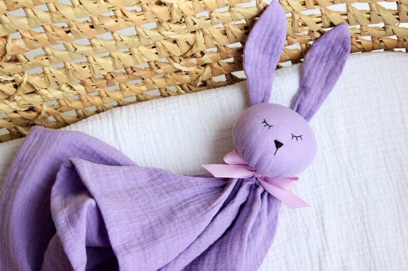 K&ouml;nnte beinhalten: Eine lavendelfarbene, bunnyf&ouml;rmige Sicherheitsdecke mit einer rosa Schleife. Die Decke ist aus weichem, strukturiertem Stoff und hat ein gesticktes Gesicht mit geschlossenen Augen.
