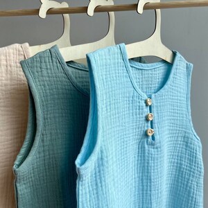 May include: Three sleeveless shirts hanging on wooden hangers. The shirts are made of a soft, textured fabric. The shirts are in shades of light pink, light blue, and light green. The light blue shirt has three wooden buttons down the front.