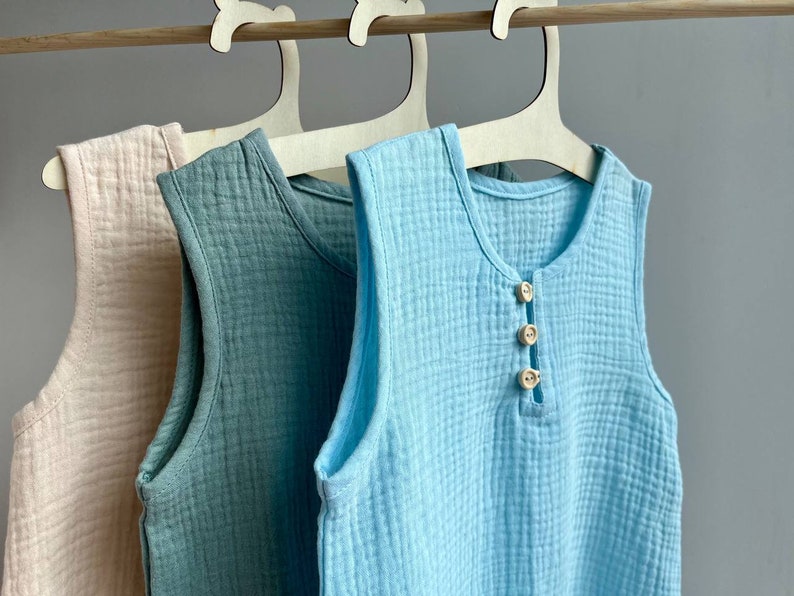 May include: Three sleeveless shirts hanging on wooden hangers. The shirts are made of a soft, textured fabric. The shirts are in shades of light pink, light blue, and light green. The light blue shirt has three wooden buttons down the front.