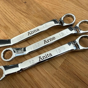 Peut inclure: Trois clés à molette chromées brillantes, gravées de noms personnalisés : "Alma", "Arne" et "Anita". Des indications de taille sont également visibles. Les clés sont posées sur un fond en bois, mettant en valeur leur finition polie et les gravures personnalisées.