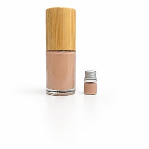 May include: A clear glass bottle of beige nail polish with a bamboo cap, next to a small glass vial of the same color. The products are set against a white background, showcasing the nail polish.