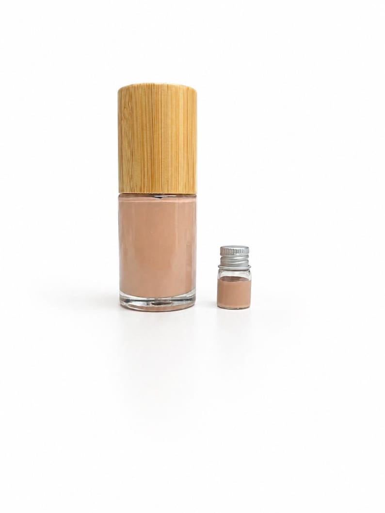 May include: A clear glass bottle of beige nail polish with a bamboo cap, next to a small glass vial of the same color. The products are set against a white background, showcasing the nail polish.