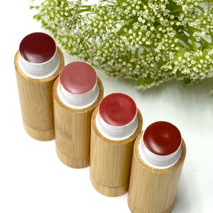 May include: Four wooden tubes with a red lip balm inside. The tubes are arranged in a row on a white surface.