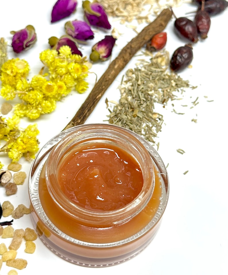 May include: A small glass jar filled with a thick, orange-coloured cream or balm. The jar is on a white surface with dried herbs and flowers scattered around it.