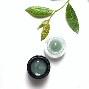 May include: Two small jars of green face cream on a white background. One jar is clear glass with a black lid, the other is black with a black lid. A green leaf branch is in the upper right corner of the image.