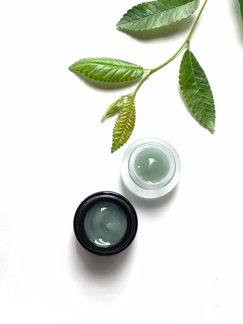 May include: Two small jars of green face cream on a white background. One jar is clear glass with a black lid, the other is black with a black lid. A green leaf branch is in the upper right corner of the image.