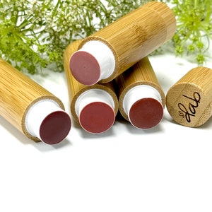May include: Four bamboo tubes with white caps and different shades of pink and red lip balm inside. The tubes are stacked on top of each other. The top tube has the word "love" engraved on the side.