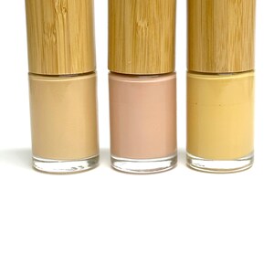 May include: Three nail polish bottles with wooden caps. The nail polish colors are beige, light pink, and yellow. The bottles are made of clear glass and are displayed against a white background.