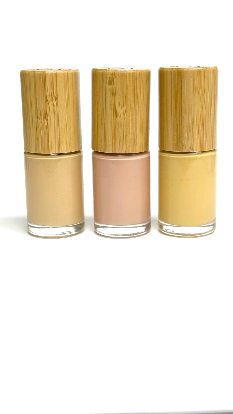 May include: Three nail polish bottles with wooden caps. The nail polish colors are beige, light pink, and yellow. The bottles are made of clear glass and are displayed against a white background.