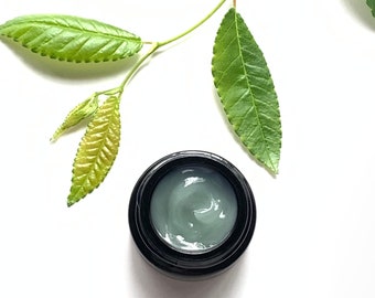 May include: A small black jar containing a green cream, beside a sprig of green leaves on a white background.