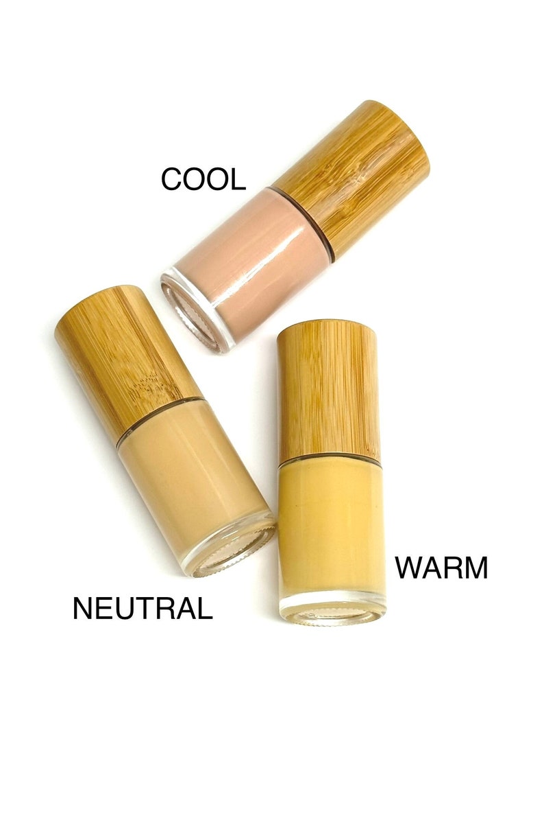 May include: Three glass bottles of foundation with bamboo lids, labeled "COOL", "NEUTRAL", and "WARM". The foundation shades range from light beige to yellow-toned, ideal for various skin tones. The bottles are on a white background.