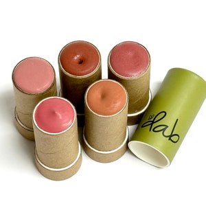 May include: Five small cardboard tubes with different shades of pink and brown lip balm inside. The tubes are arranged in a circle with a green cardboard tube with the word "dab" written on it in black.