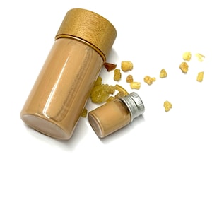 May include: Two clear glass bottles, one large and one small, filled with a light brown liquid. The larger bottle has a bamboo lid with a gold band. Scattered around are small, amber-colored resin pieces. The background is white.