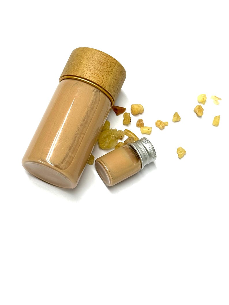 May include: Two clear glass bottles, one large and one small, filled with a light brown liquid. The larger bottle has a bamboo lid with a gold band. Scattered around are small, amber-colored resin pieces. The background is white.