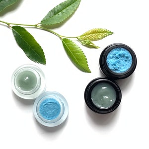May include: Three small jars of face cream, two with a light green cream and one with a light blue cream. The jars are on a white background with a green leafy branch.