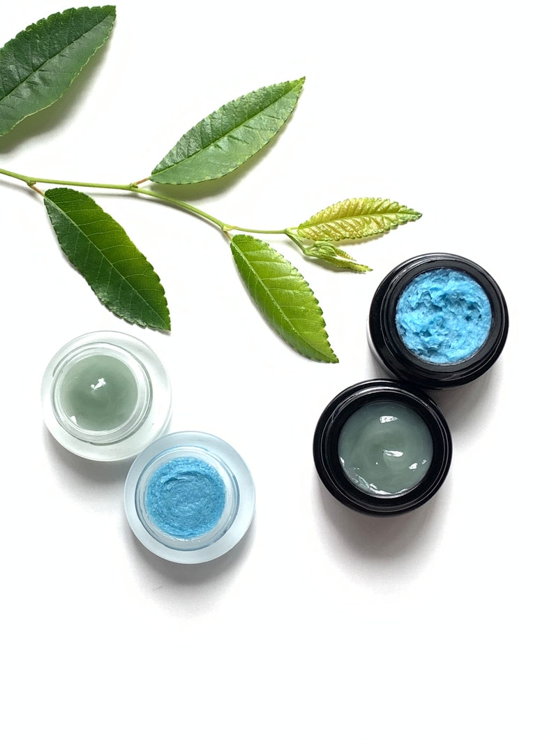 May include: Three small jars of face cream, two with a light green cream and one with a light blue cream. The jars are on a white background with a green leafy branch.