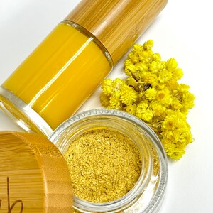 Pu&ograve; includere: Un barattolo di vetro con un coperchio dorato e una polvere gialla all'interno. Il barattolo &egrave; appoggiato su una superficie bianca accanto a un piccolo mazzo di fiori gialli e una bottiglia di vetro con un liquido giallo e un tappo di legno.