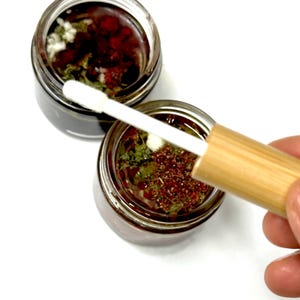 May include: Two small clear glass jars filled with a dark liquid containing visible green herbs and small red particles. A wooden-handled applicator with a white sponge tip is positioned over the open jars, showing the texture of the contents.
