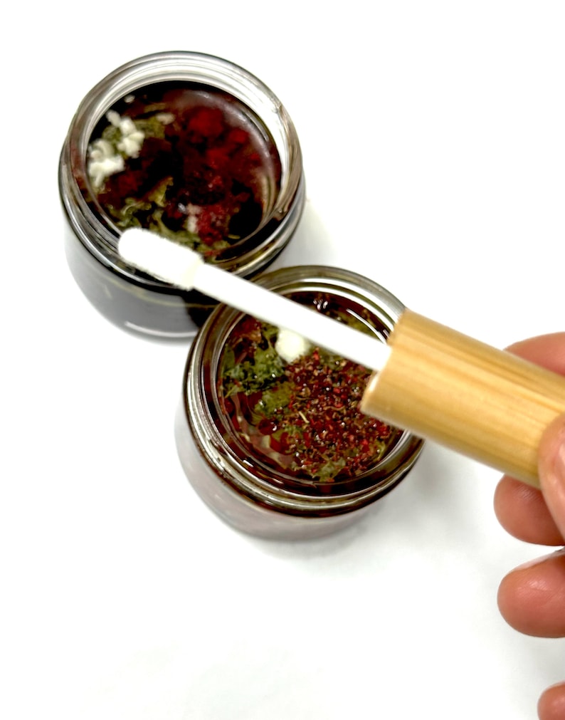 May include: Two small clear glass jars filled with a dark liquid containing visible green herbs and small red particles. A wooden-handled applicator with a white sponge tip is positioned over the open jars, showing the texture of the contents.