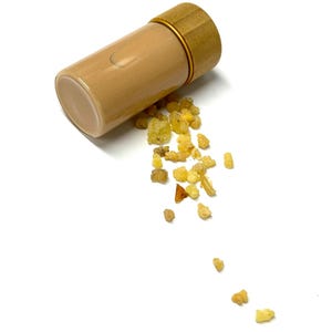May include: A tan glass vial with a bamboo lid and gold-colored ring, lying on its side with small, amber-colored resin pieces spilling out. The vial is cylindrical and appears to be empty. The background is plain white.