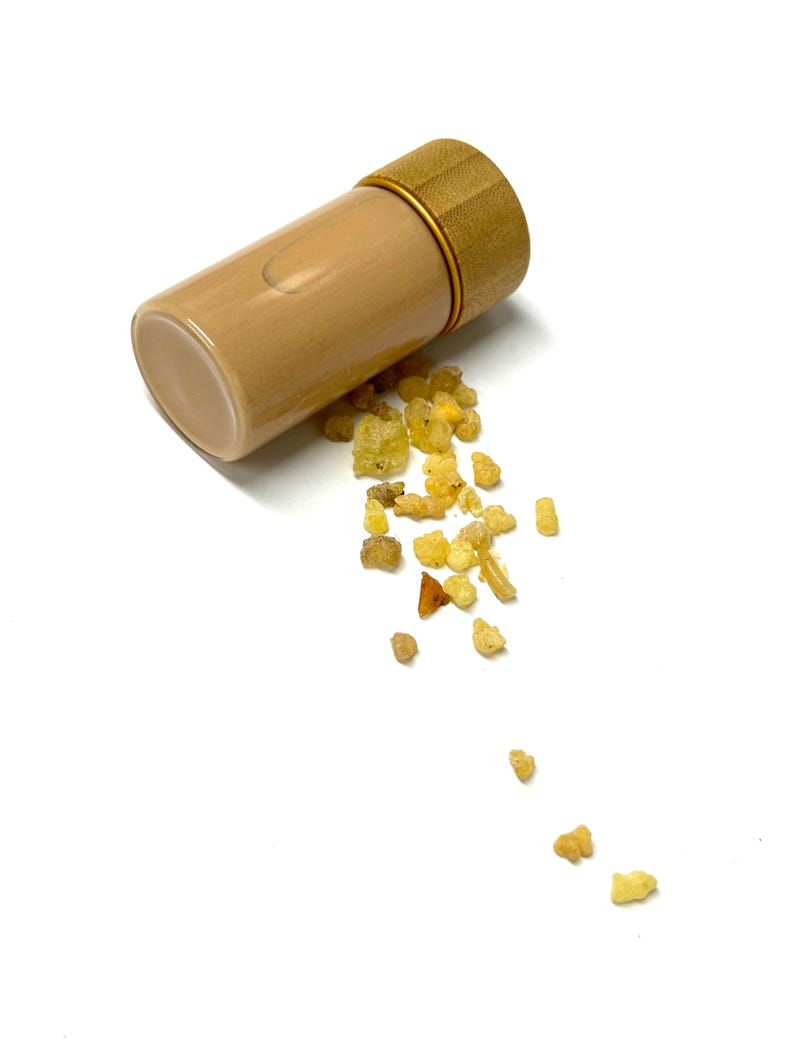 May include: A tan glass vial with a bamboo lid and gold-colored ring, lying on its side with small, amber-colored resin pieces spilling out. The vial is cylindrical and appears to be empty. The background is plain white.