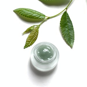 May include: A small glass jar with a frosted lid containing a green cream. A sprig of green leaves is placed on a white background.