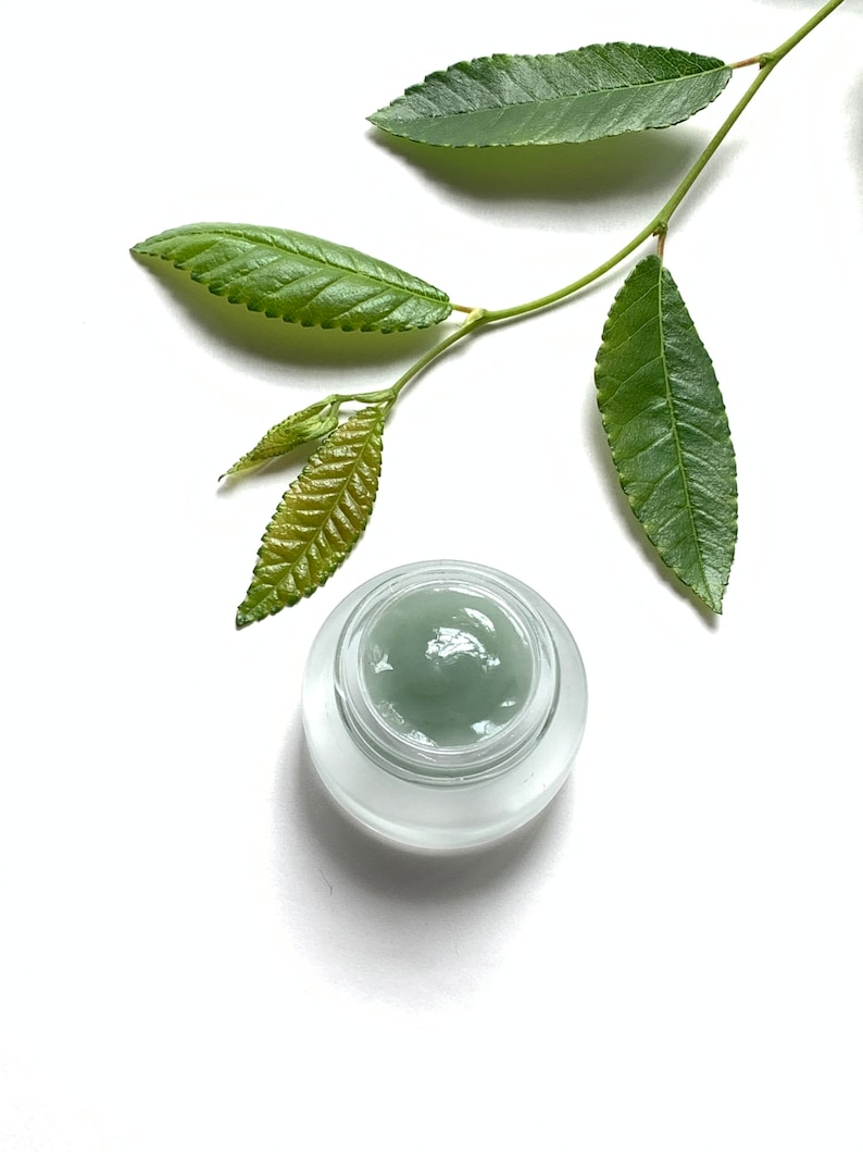 May include: A small glass jar with a frosted lid containing a green cream. A sprig of green leaves is placed on a white background.
