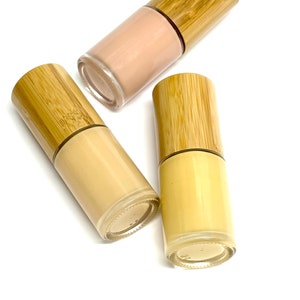 May include: Three glass bottles of foundation with bamboo lids. The foundation shades vary from light beige to a deeper tan color. The bottles are arranged on a white background.