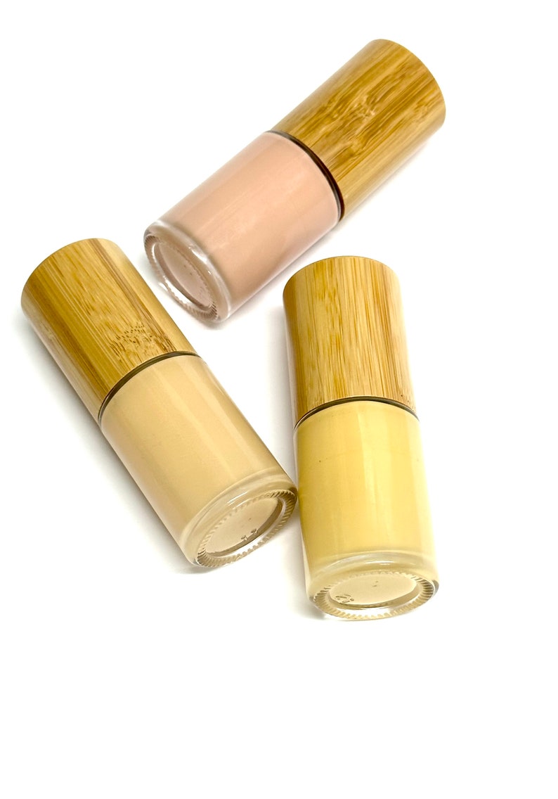 May include: Three glass bottles of foundation with bamboo lids. The foundation shades vary from light beige to a deeper tan color. The bottles are arranged on a white background.