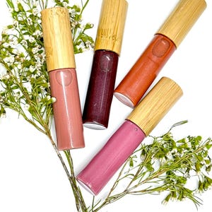 May include: Four cylindrical lip gloss tubes with light-colored bamboo caps, each containing a different shade of liquid cosmetic: a soft dusty rose, a deep dark berry, a warm terracotta orange, and a vibrant mauve pink, arranged with white flowers.