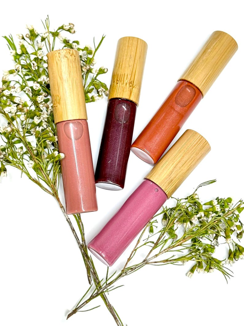 May include: Four cylindrical lip gloss tubes with light-colored bamboo caps, each containing a different shade of liquid cosmetic: a soft dusty rose, a deep dark berry, a warm terracotta orange, and a vibrant mauve pink, arranged with white flowers.