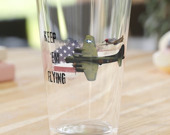 May include: A clear glass with a patriotic design featuring an American flag and a vintage airplane. The text "KEEP 'EM FLYING" is printed on the glass. The glass is sitting on a wooden table.