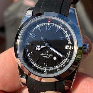 May include: A black and silver wristwatch with a black rubber band. The watch face has white markings and a small white square with the text 'Automatic 20BAR'.
