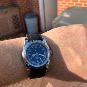 May include: A stainless steel wristwatch with a black dial and a black rubber strap. The watch face features white numerals and hands, with a date display at the 3 o'clock position. The watch is on a wrist.