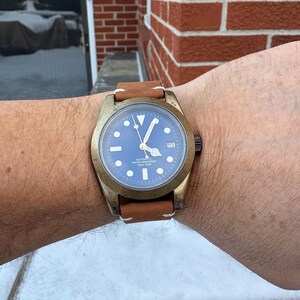 Pu&ograve; includere: Un orologio da polso color bronzo con quadrante blu e cinturino in pelle marrone. L'orologio ha indici e lancette bianche e un datario a ore 3. Il cinturino &egrave; cucito con filo bianco.