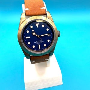 Pu&ograve; includere: Un orologio da polso color bronzo con quadrante blu navy e cinturino in pelle marrone. L'orologio ha indici bianchi e un datario a ore 3. Il testo "AUTOMATIC WATER RESISTANT 100m: 330ft" &egrave; stampato sul quadrante.
