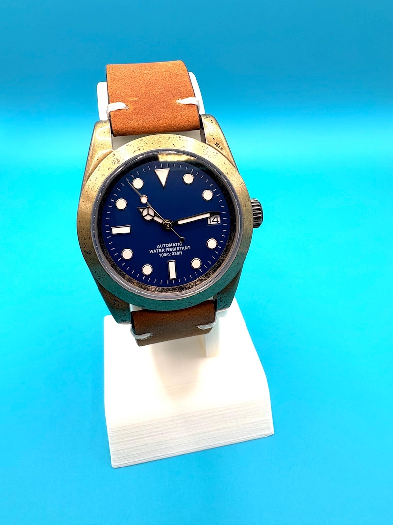 Pu&ograve; includere: Un orologio da polso color bronzo con quadrante blu navy e cinturino in pelle marrone. L'orologio ha indici bianchi e un datario a ore 3. Il testo "AUTOMATIC WATER RESISTANT 100m: 330ft" &egrave; stampato sul quadrante.