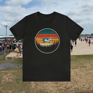 May include: Black t-shirt with a vintage-style graphic of a small airplane flying over a forest with a sunset in the background.
