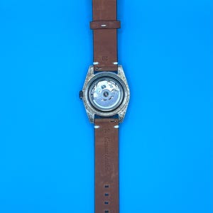 Pu&ograve; includere: Un cinturino in pelle marrone con un quadrante argentato. Il quadrante ha un retro trasparente, che rivela il meccanismo interno. Il cinturino &egrave; etichettato "Genuine Leather". Lo sfondo &egrave; blu.