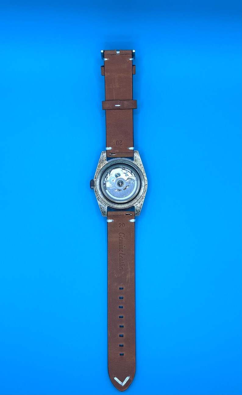 Pu&ograve; includere: Un cinturino in pelle marrone con un quadrante argentato. Il quadrante ha un retro trasparente, che rivela il meccanismo interno. Il cinturino &egrave; etichettato "Genuine Leather". Lo sfondo &egrave; blu.