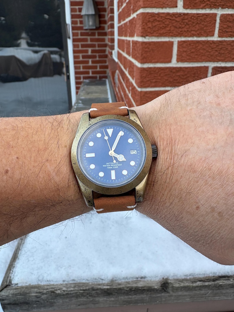May include: A bronze-colored wristwatch with a blue face and a brown leather strap. The watch has white hour markers and hands, and a date display at the 3 o'clock position. The strap is stitched with white thread.