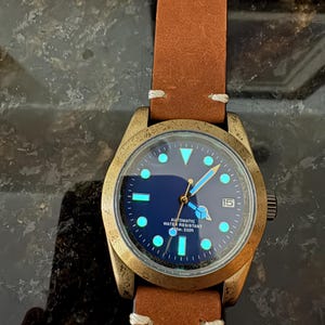 Pu&ograve; includere: Un orologio da polso color bronzo con cinturino in pelle marrone. Il quadrante &egrave; blu scuro con indici e lancette luminose. Il testo "AUTOMATIC WATER RESISTANT 100m 330ft" &egrave; visibile. Visualizzazione della data.