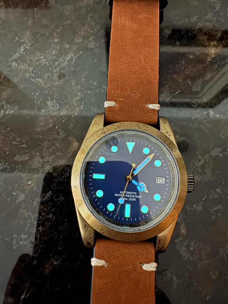 Pu&ograve; includere: Un orologio da polso color bronzo con cinturino in pelle marrone. Il quadrante &egrave; blu scuro con indici e lancette luminose. Il testo "AUTOMATIC WATER RESISTANT 100m 330ft" &egrave; visibile. Visualizzazione della data.