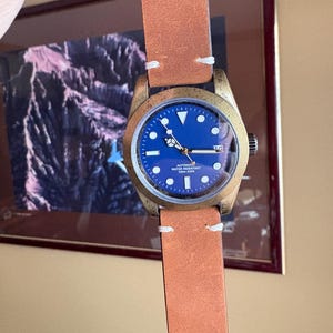 Pu&ograve; includere: Un orologio da polso color bronzo con quadrante blu e cinturino in pelle marrone. L'orologio ha cuciture bianche sul cinturino e un datario. Il quadrante ha indici e lancette bianche.