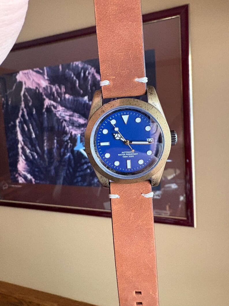 Pu&ograve; includere: Un orologio da polso color bronzo con quadrante blu e cinturino in pelle marrone. L'orologio ha cuciture bianche sul cinturino e un datario. Il quadrante ha indici e lancette bianche.