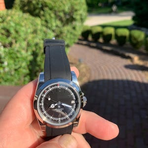 May include: A stainless steel wristwatch with a black dial and a black rubber strap. The watch features a date window and luminous hands. The watch is held in a hand against a blurred background of greenery.