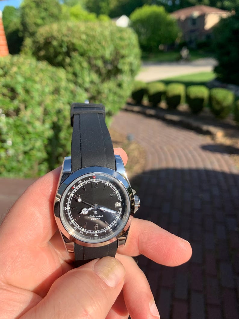 May include: A stainless steel wristwatch with a black dial and a black rubber strap. The watch features a date window and luminous hands. The watch is held in a hand against a blurred background of greenery.